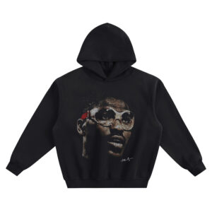Hakeem Olajuwon Big Face Hoodie graphic showing a close-up portrait of Hakeem Olajuwon wearing oversized clear goggles with a red strap detail and a small signature-style mark on a black hoodie.