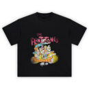 The Flintstones Graphic Tee graphic showing pink “THE FLINTSTONES” title above a crowded stone-age car scene with multiple cartoon characters riding together and dust puffs below on a black tee.